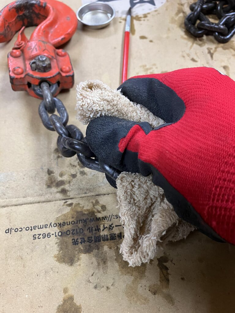 レバーホイストのチェーンに付着したラストリムーバーをウエスで拭き取るメンテナンス作業の様子。